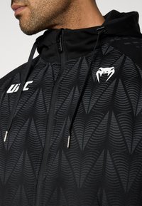 Sweat à capuche noir zippé avec un motif ondulé texturé, des cordons de serrage et un logo blanc sur la poitrine. Tissu doux avec une coupe ajustée.