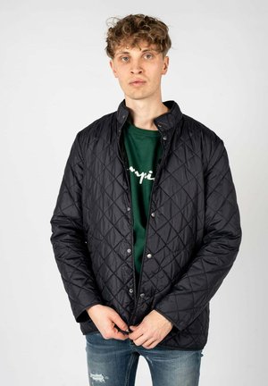 Kurtka pikowana w kolorze czarnym z zatrzaskami, z wysokim kołnierzem i bocznymi kieszeniami, noszona na zielonym t-shircie i w połączeniu z przetartymi dżinsami.
