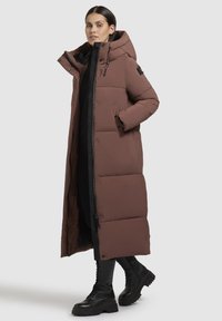 Hosszú barna pufferekőkabát kapucnival, steppelt részekkel, elülső cipzárral és patentgombokkal. Fekete fűzős csizmával viselve.