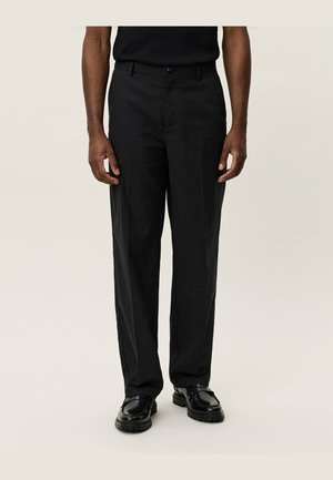 Homme portant un pantalon noir droit, des chaussures en cuir noires et une chemise noire à manches courtes sur un fond uni.