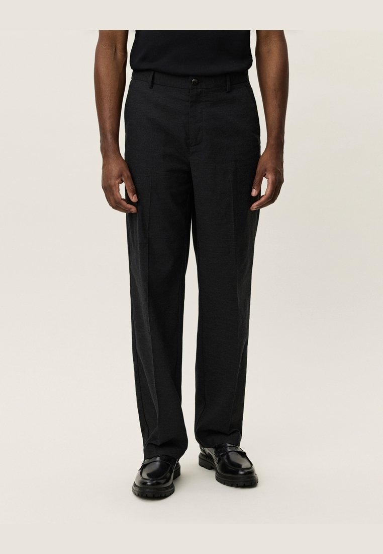 Homme portant un pantalon noir droit, des chaussures en cuir noires et une chemise noire à manches courtes sur un fond uni.