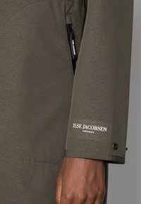 Zielona wodoodporna kurtka z teksturowaną tkaniną. Posiada kieszeń na zamek błyskawiczny i białe logo "ILSE JACOBSEN HORNBÆK" na mankiecie.