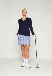 Μπλέ ριγέ πλεκτό πουλόβερ με V λαιμόκοψη; συνδυασμένο με μίνι φούστα σε μπλε λεοπάρ σχέδιο. Το μοντέλο κρατάει ένα μπαλάκι του γκολφ, φορώντας λευκές κάλτσες και παπούτσια.