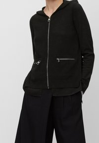 Black zip-up hoodie made of soft fabric, featuring a front zipper and two zippered pockets at the waist. Hooded design with a relaxed fit.