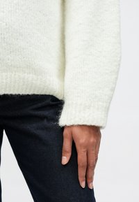 Hvit strikket genser med ribbet kant og mansjetter, som viser en myk, lodden tekstur, kombinert med mørke denimjeans.