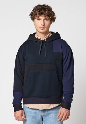 Jeune homme aux cheveux bouclés portant un sweat à capuche color-block bleu marine avec un logo d'arbre et un jean bleu clair, debout devant un fond uni.
