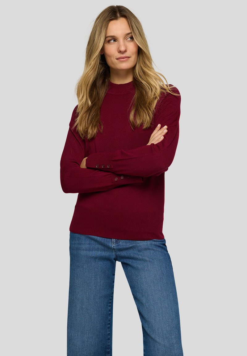 Weinroter Strickpullover mit langen Ärmeln, geripptem Kragen und Knopfdetails an den Bündchen, kombiniert mit blauen Denim-Jeans.