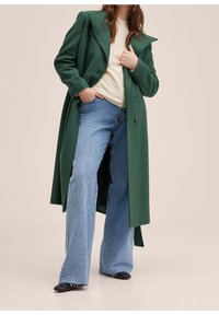 Grøn lang frakke med store revers, dobbeltradet front, båret over en cremet sweater og blå vidde jeans, kombineret med sorte sko.