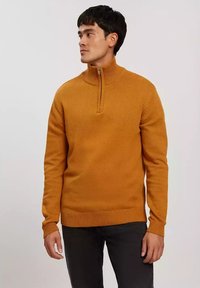 Pomarańczowy sweter z dzianiny z wysokim kołnierzem i zamkiem błyskawicznym na ćwiartkę, z prążkowanymi mankietami i dolnym brzegiem, noszony na ciemnych dżinsach.