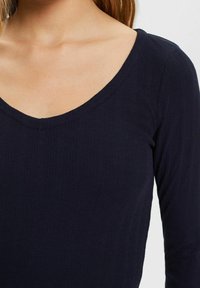 Mörkblå långärmad topp med V-ringning, som har ribbat mönster och en åtsittande design. Tyget verkar mjukt och elastiskt.