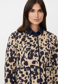 Giacca trapuntata con base beige e stampa leopardata blu navy. Presenta un colletto in denim, tasche frontali e piping a contrasto lungo i bordi.