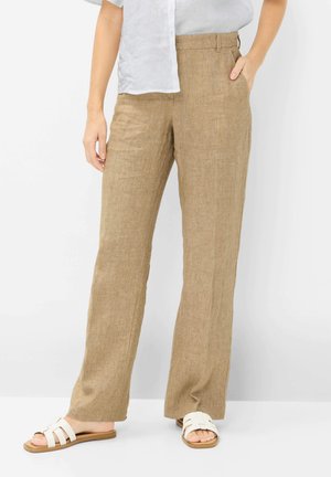 Femme portant un pantalon en lin beige avec poches et des sandales blanches à enfiler, debout devant un fond blanc uni.
