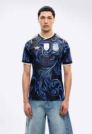 Joven con camiseta de fútbol negra con patrones azules en espiral, insignia de la FIFA 2022 y escudo de la AFA, combinado con jeans azul claro.