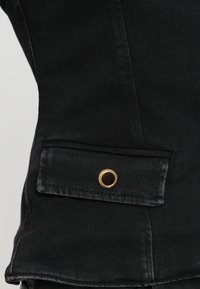 Detail van een zwarte denimjack met een voorkantzak met een gouden drukknoop en zichtbare stiksels. De textuur lijkt glad en mat.