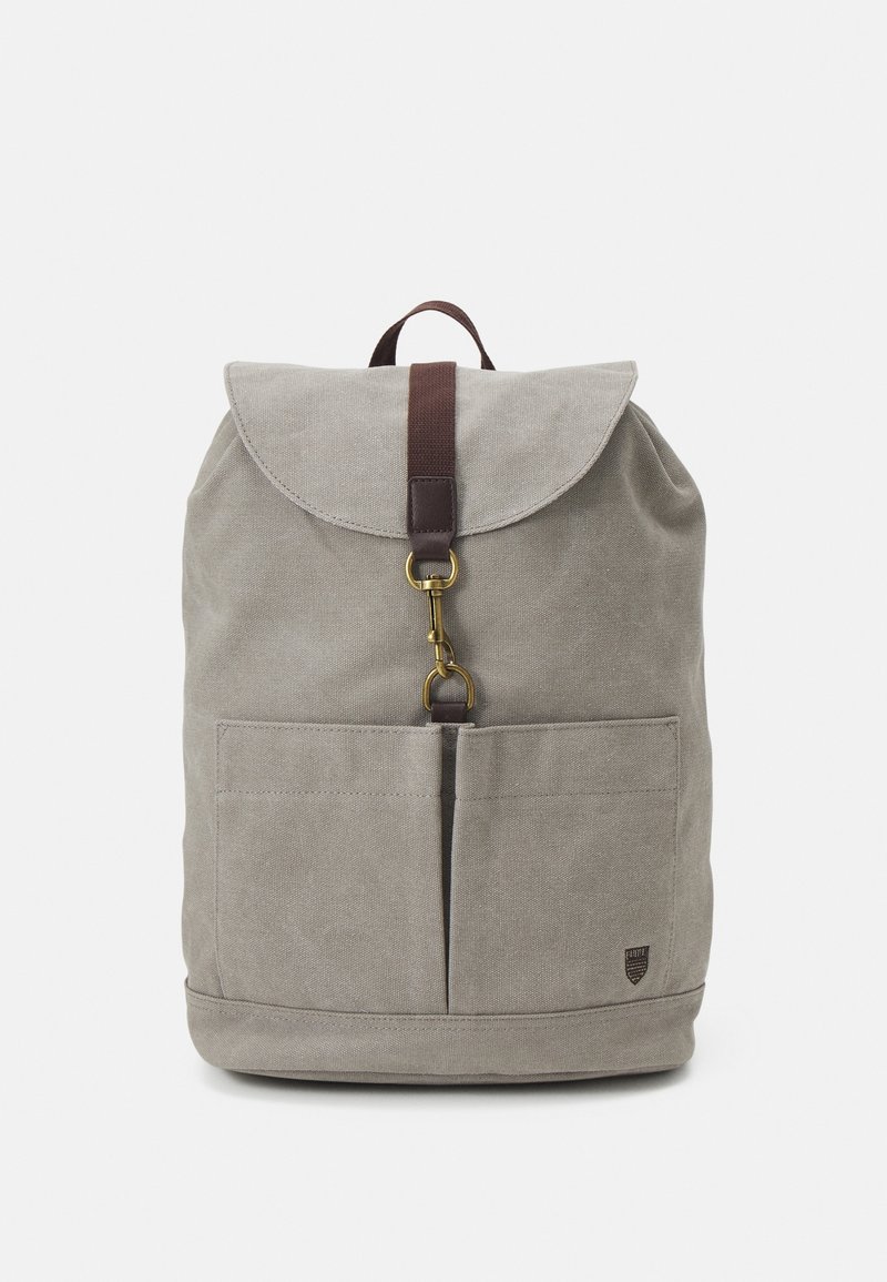 Sac à dos en toile gris clair avec rabat avant maintenu par une sangle marron et une attache en laiton, comprenant deux poches frontales et une petite étiquette avec logo.