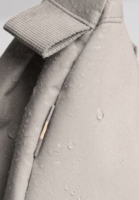 Tessuto grigio resistente all'acqua con una superficie testurizzata, dotato di un cappuccio superiore e un piccolo accento metallico. Gocce visibili sul materiale.