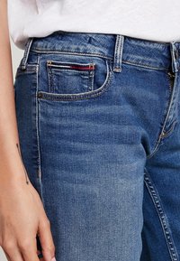 Modré džínové kalhoty s klasickým pětipocketovým designem, na látce s opotřebením a malým akcentem červené a bílé barvy v kapsičce na mince.