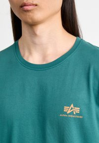 Turkusowa bawełniana koszulka z okrągłym dekoltem i krótkimi rękawami. Posiada pomarańczowe logo "Alpha Industries" po lewej stronie. Gładka struktura.