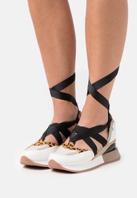 Sportovní boty s bílým a béžovým základem, akcenty s leopardím potiskem a černými textilními pásky křížícími se kolem kotníků. Gumová podrážka.