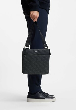 Sort crossbody taske med et struktureret mønster, der har en frontlomme med lynlås og justerbar rem, holdt af en person iført mørkt tøj.