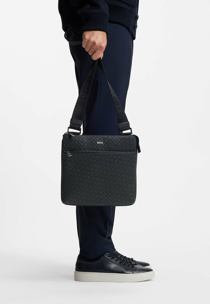 Borsa a tracolla nera con un motivo testurizzato, dotata di una tasca frontale con zip e tracolla regolabile, tenuta da una persona in abbigliamento scuro.