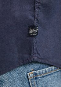 Tummansininen kangas, jossa on teksturoitu pinta; siinä on "Petrol Industries" -merkki ommeltuna saumaan; yhdistetty vaaleansinisiin farkkuihin.