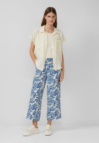 Lichtgele kortekorte shirt met twee zakken, gecombineerd met blauwe en witte geblokte wijde broek. Model draagt off-white sneakers.