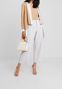 Beige Strickjacke mit weißer Vorderseite, heller karierter Culotte-Hose, beige Riemchensandalen und einer weißen Handtasche mit Holzgriff.