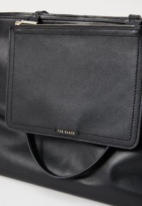 Zwarte leren tote bag met een gestructureerd oppervlak, een voorzak en gouden hardware. Beschikt over een prominent logo-label en gladde handvatten.