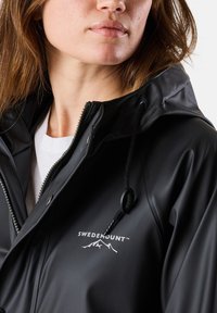 Schwarze wasserdichte Jacke mit Reißverschluss und Kapuze. Merkmale ein Logo "SWEDEMOUNT" in Weiß. Glatte Textur, verstellbare Kordeln.