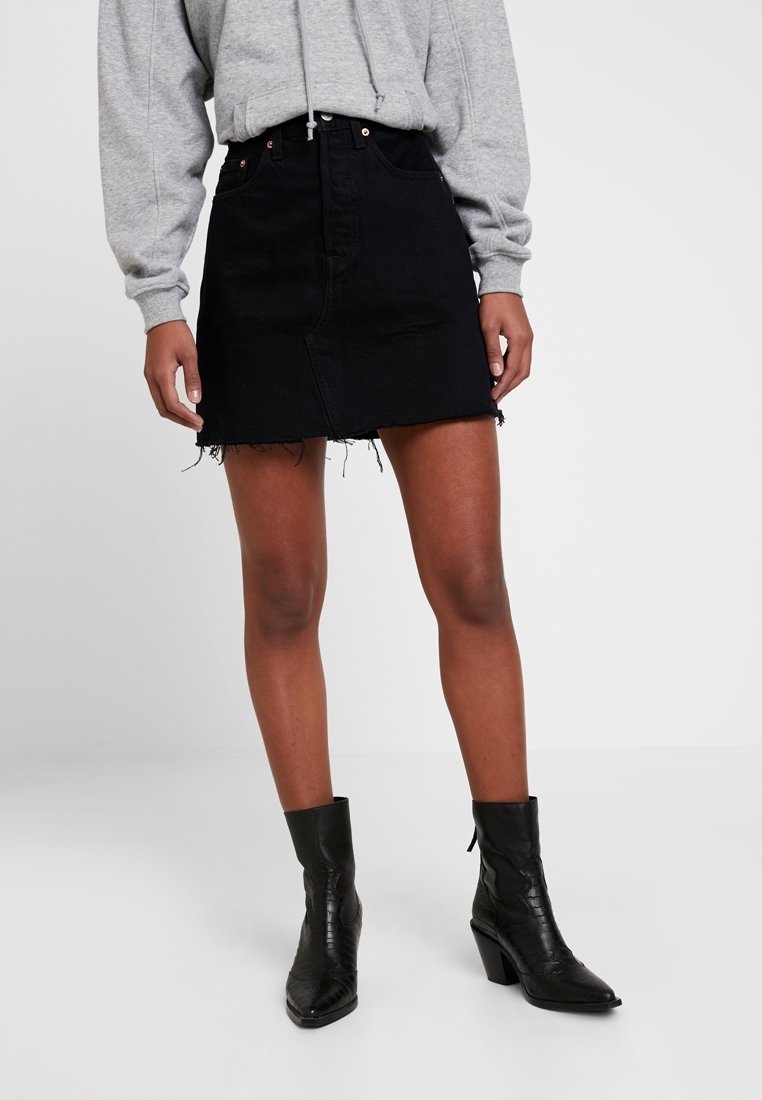 Femme portant un sweat à capuche gris, une mini-jupe en jean noire déchirée et des bottines noires à talons, debout sur un fond clair uni.