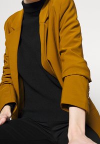 Personne portant un blazer jaune moutarde sur un pull à col roulé côtelé noir et un pantalon noir, assise avec un bras reposant sur le genou.