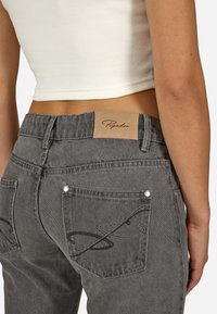 Zbliżenie na osobę w szarych dżinsach z skórzaną naszywką marki oraz haftowanym wzorem na tylnej kieszeni, połączonych z białym krótkim topem.