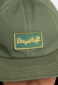 Casquette verte avec des détails cousus et une étiquette rectangulaire. L'étiquette porte "Dayshift" en doré en cursive sur un fond teal.