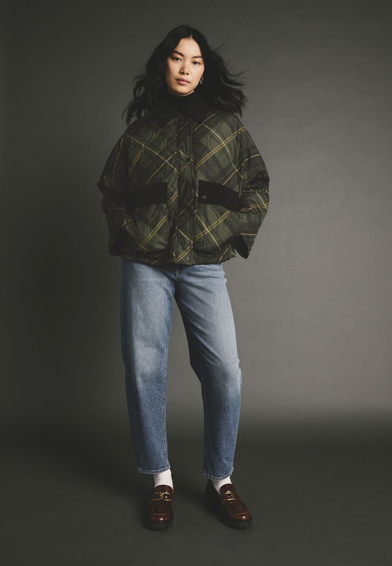 Gequilted groene en zwarte geruite pufferjack met zwarte accenten, gecombineerd met lichtblauwe rechtomslagjeans en bruine instapschoenen.