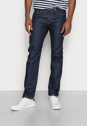 Mann trägt dunkelblaue Slim-Fit-Jeans, weiße Sneakers und ein schwarz-weiß gestreiftes Hemd und steht vor einem neutralen Hintergrund.