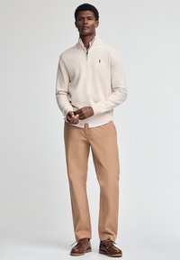Maglione beige con zip e polsini a costine, abbinato a pantaloni marrone chiaro e scarpe marroni. L'outfit presenta un logo sul maglione.
