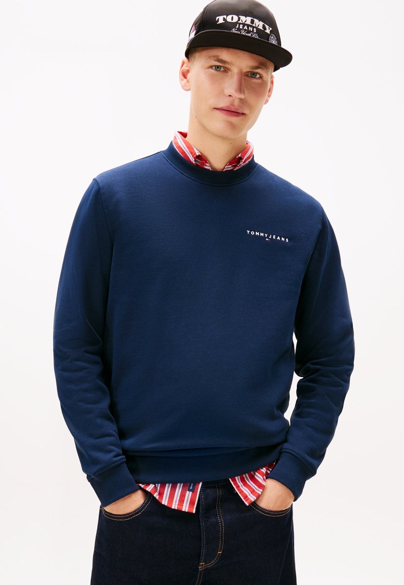 Νέος άνδρας που φοράει μπλε σκούρο φούτερ Tommy Jeans, από κάτω μια κόκκινη και λευκή ριγέ πουκάμισο, σκούρο τζιν και μαύρο καπέλο Tommy Jeans.