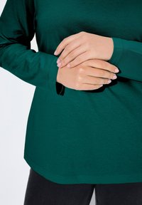 Zelená dlouhá rukávová košile vyrobená z měkké tkaniny, s kulatým lemem a volným střihem; zahrnuje rozparky na zápěstích pro větší pohodlí.