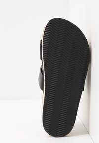 Semelle de sandale noire avec motif de bande en chevron inclinée contre un coin de mur et de sol blancs.