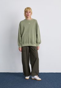 Personne debout portant un sweat-shirt Adidas vert clair, un pantalon cargo ample vert foncé et des baskets blanches avec des rayures roses sur un tapis bleu.