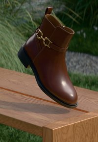 Braune Leder-Ankle-Boots mit goldener Kettenakzent, abgerundeter Zehenpartie, glatter Textur, bescheidenem Absatz und einem Knöchelriemen. Auf einer Holzoberfläche platziert.