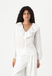 Blusa bianca a maniche lunghe con colletto arricciato, scollo a V e chiusura con bottoni. Realizzata in un tessuto leggero e testurizzato.