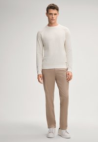 Hellbeige strukturierter Pullover, cremefarbene Hose und weiße Sneaker. Model steht direkt zur Kamera, vor einem neutralen Hintergrund.
