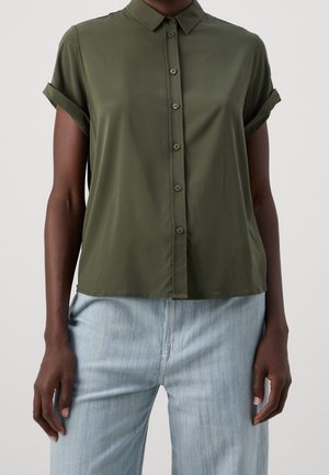 Personne portant une chemise boutonnée à manches courtes de couleur vert olive avec les poignets retroussés et un jean taille haute bleu clair, sur un fond gris.
