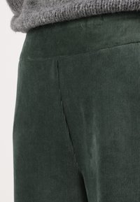Pantaloni in corduroy verde con texture a righe verticali. Presentano una vita aderente e un sottile dettaglio delle cuciture sul retro. Materiale morbido e soffice.