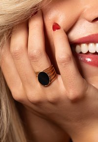 Goudkleurige ring met vijf gestapelde banden, met in het midden een ovale zwarte steen. Glad oppervlak, gepolijste afwerking en subtiele details.