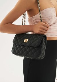 Femme portant un haut rose froncé et une jupe noire tenant un sac à épaule matelassé noir avec des anses en chaîne dorée et un fermoir.