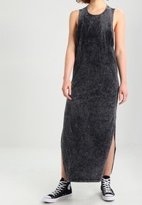 Kobieta w bezrękawnikowej, ciemnoszarej sukience do kostek z bocznymi rozporkami oraz czarnych wysokich trampkach z białymi sznurówkami.