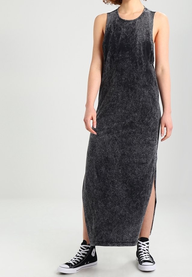 Kobieta w bezrękawnikowej, ciemnoszarej sukience do kostek z bocznymi rozporkami oraz czarnych wysokich trampkach z białymi sznurówkami.